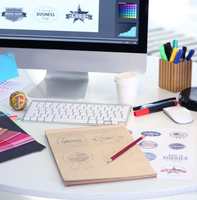 Creative workspace with logo sketches, color swatches, and computer showing digital logo designs.