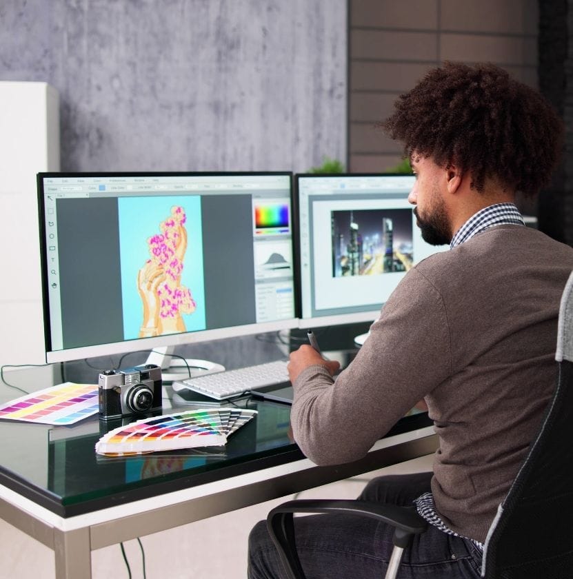 Male graphic designer creating digital art on dual monitors with color swatches and a camera on desk.
