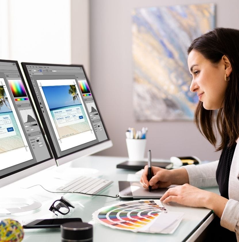 Female graphic designer working on a website layout using dual monitors and a color palette.