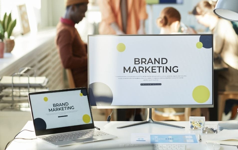 Dual screens displaying “Brand Marketing” in a bright creative office environment with people working in the background.